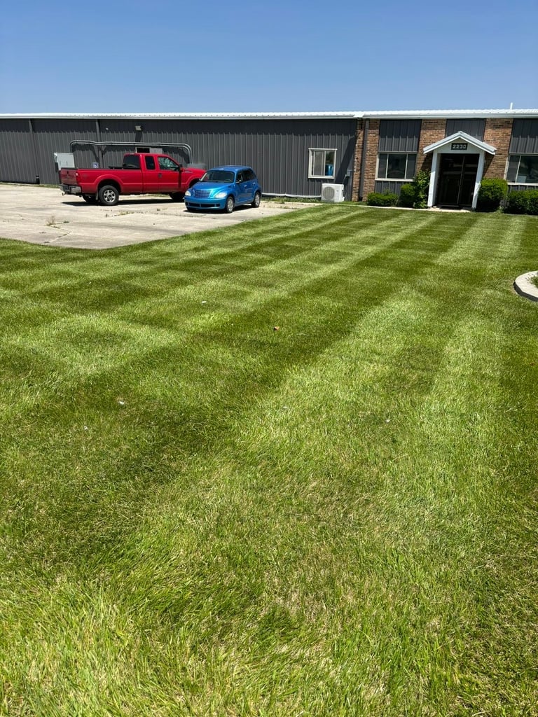 A red pickup truck and blue car parked on a concrete pad in front of a large industrial building with manicured green lawn in foreground