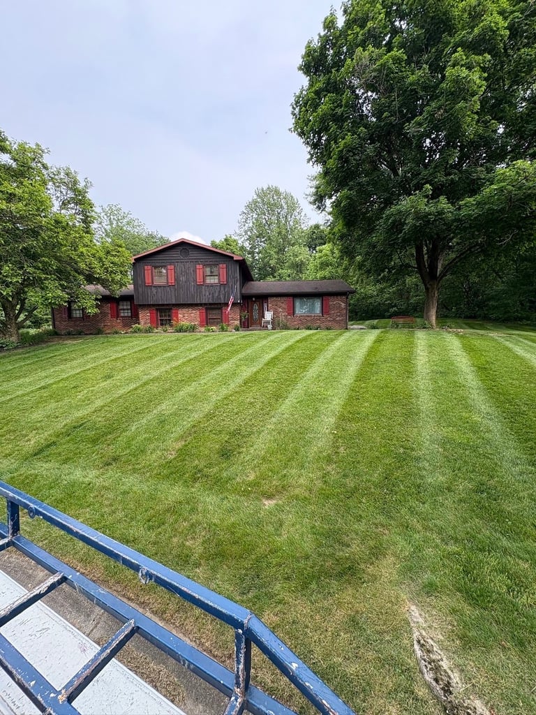 Red barn with striped mowed lawn and blue metal fence in foreground, surrounded by green trees