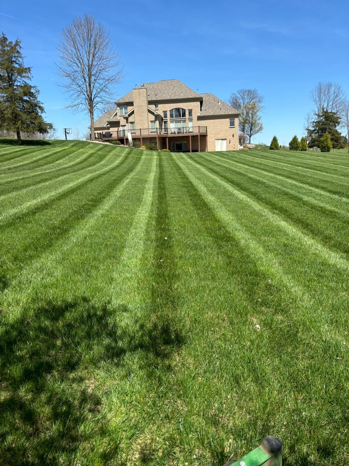 Large tan brick house with manicured lawn striped with mowing patterns, surrounded by trees under clear blue sky
