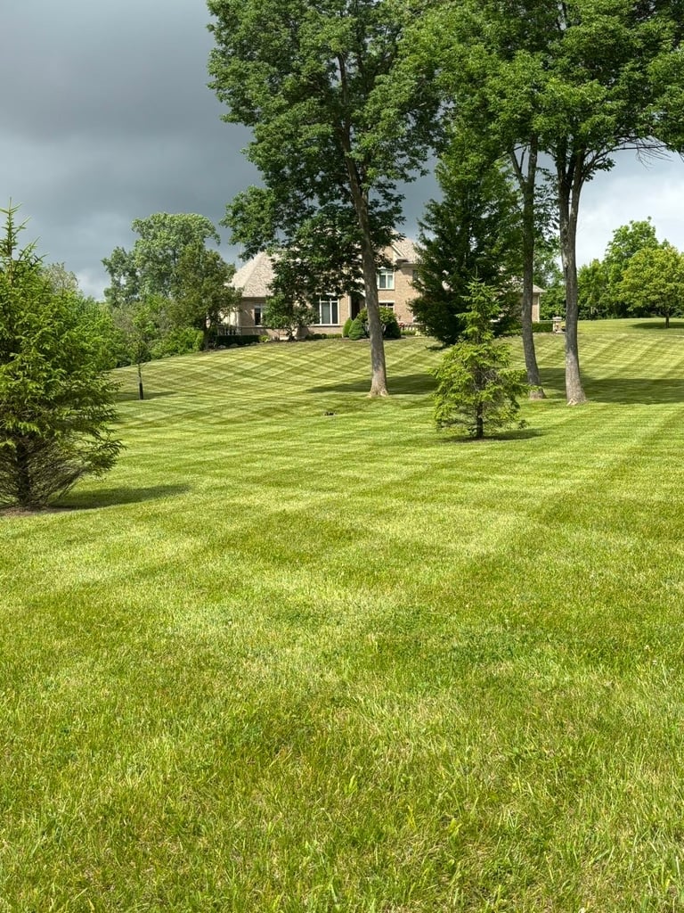 Large brick house set on expansive manicured lawn with mature trees under cloudy sky